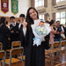 A smiling graduate in a black gown stands in a school hall holding a graduation bouquet of white rose and light blue baby's breath with a graduation teddy bear. 