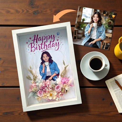 A white shadow box frame on a wooden table, featuring a custom illustration of a woman with 'Happy Birthday' text and pink flowers. A small photo of the woman is shown in the top right with an arrow pointing to the artwork.