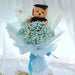A graduation-themed flower bouquet featuring a small teddy bear wearing a black graduation cap and gown, surrounded by pale blue baby's breath flowers and wrapped in light blue tissue paper with a blue ribbon. 
