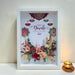 A framed decorative wall art piece with a white frame and a printed Diwali design featuring the words 'Happy Diwali Festival of Lights,' colorful mandala motifs, hanging diyas, and a floral arrangement at the bottom. It is photographed leaning against a textured off-white wall on a white surface, with a small lit gold cup on a saucer to the right.