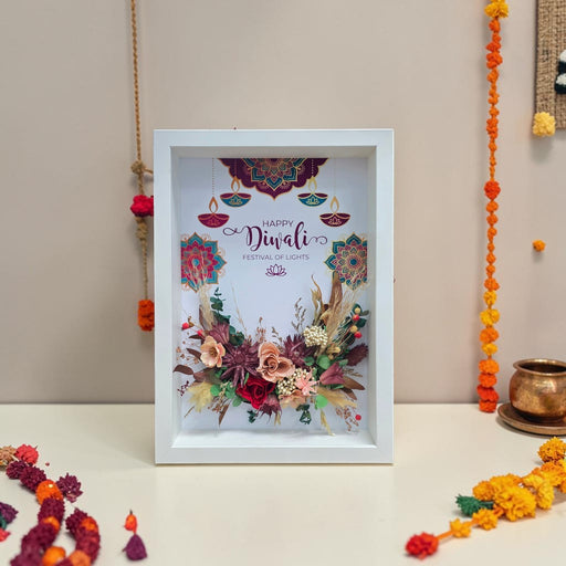 A framed Diwali art print featuring a white background with colorful mandala designs, hanging diyas, and a floral arrangement at the bottom. The frame is displayed on a white surface against a beige wall, surrounded by festive garlands and a brass pot.