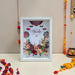 A framed Diwali art print featuring a white background with colorful mandala designs, hanging diyas, and a floral arrangement at the bottom. The frame is displayed on a white surface against a beige wall, surrounded by festive garlands and a brass pot.