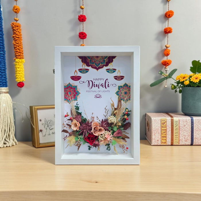 A white square shadow box frame displaying a colorful Diwali-themed floral and mandala design with the words 'Happy Diwali Festival of Lights,' placed on a wooden shelf in a decorated indoor setting.