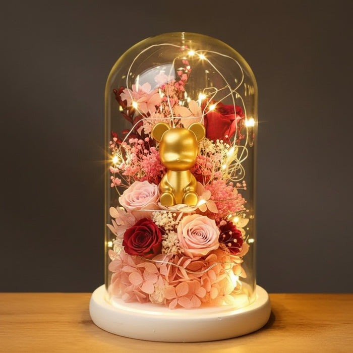 A decorative glass dome display on a white round base, featuring a small gold bear figurine surrounded by pink and red roses, dried flowers, and warm string lights against a dark gray background.