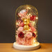 A decorative glass dome display on a white round base, featuring a small gold bear figurine surrounded by pink and red roses, dried flowers, and warm string lights against a dark gray background.