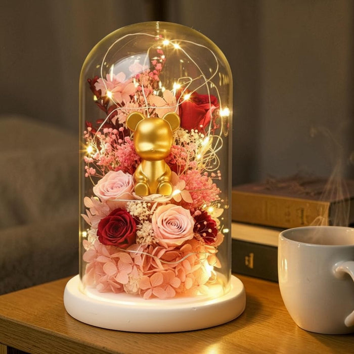 A decorative glass dome lamp with a small gold bear figurine, pink and red roses, and warm fairy lights, displayed on a wooden table next to a white mug in a cozy indoor setting.