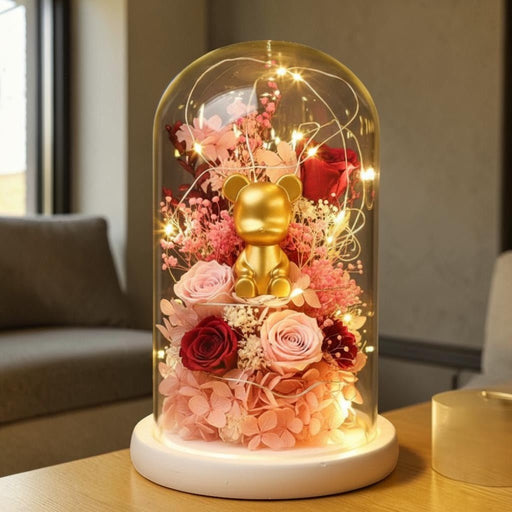 A decorative glass cloche dome on a white round base, containing a small gold bear figurine surrounded by pink and red preserved flowers and warm string lights, placed on a table in a softly lit indoor setting.
