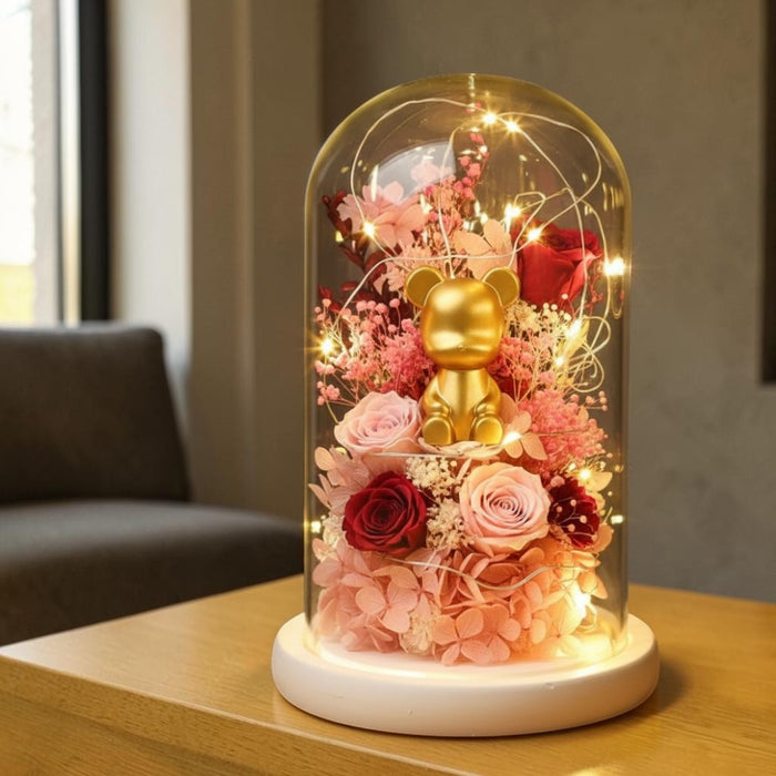A decorative glass cloche dome lamp with a small gold bear figurine, surrounded by pink and red roses, dried flowers, and warm string lights, displayed on a wooden table in a softly blurred indoor setting.