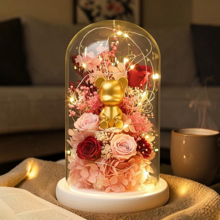 A glass dome flower display with a small gold bear figurine, pink and red roses, baby's breath, and warm string lights on a white base, placed on a table in a cozy indoor setting.