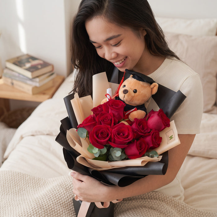 A woman smiling while holding a bouquet of red roses wrapped in black paper, with a small teddy bear wearing a graduation cap nestled inside