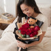 A woman smiling while holding a bouquet of red roses wrapped in black paper, with a small teddy bear wearing a graduation cap nestled inside