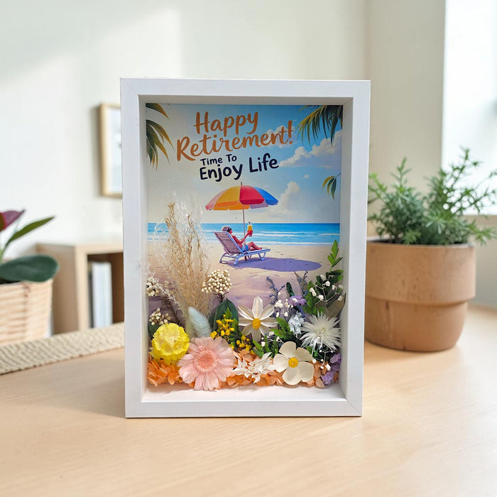 A framed retirement shadow box card standing on a light wood table. The white frame contains a colorful beach scene with palm leaves, flowers, and the text 'Happy Retirement! Time To Enjoy Life'.