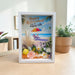 A framed retirement shadow box card standing on a light wood table. The white frame contains a colorful beach scene with palm leaves, flowers, and the text 'Happy Retirement! Time To Enjoy Life'.